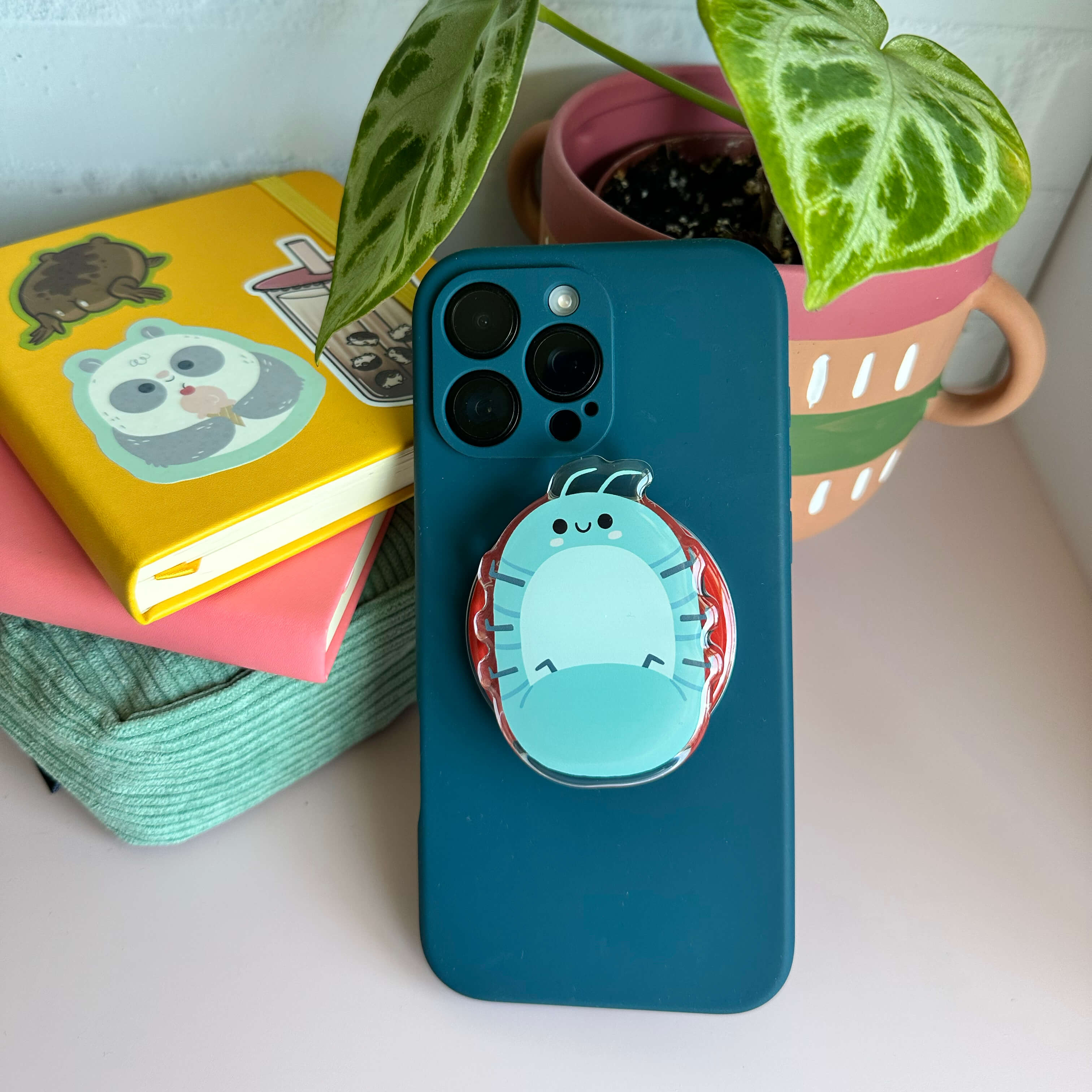 Blue phone case with a magnetic phone grip of a roly poly on a white surface with books and a plant.