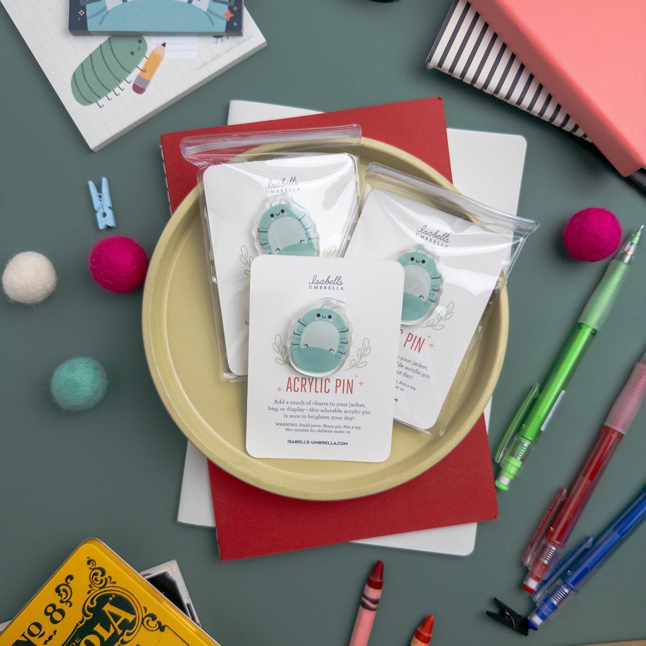 Three cute Roly Poly acrylic pin with glossy epoxy finish packaged and set on a yellow plate surrounded by a variety of art supplies.