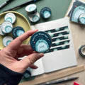 A hand holding a roll of Roly Poly-themed Washi tape with a dark background, featuring a smiling isopod illustration. More rolls of the same Washi tape and an opened journal decorated with strips of the tape are visible in the background.
