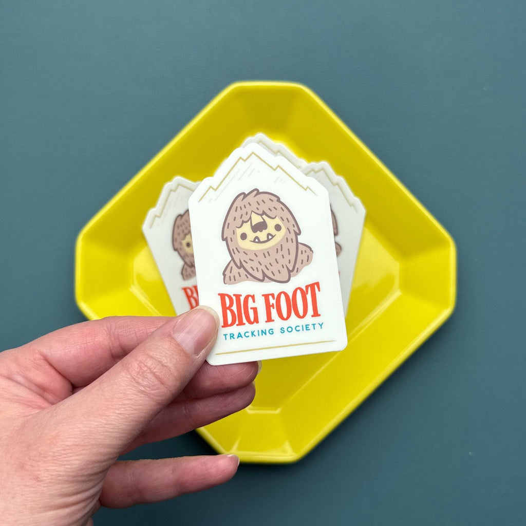 Hand holding a sticker of a big foot with mountains in the background