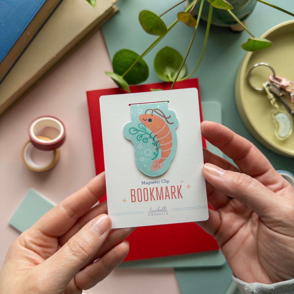 Hand holding a shrimp bookmark with a magnetic clip design on a desk with stationery items.