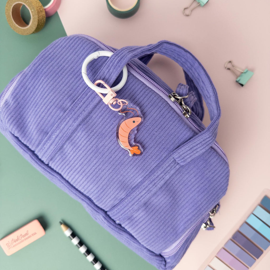 Purple pencil case with a Shrimp acrylic charm keychain attached on a colorful background with stationery items