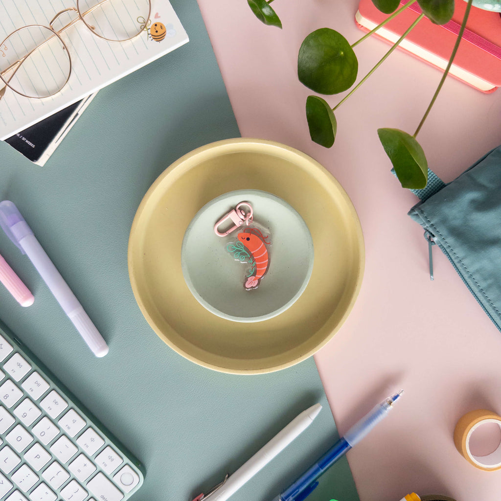 Yellow dish with a pink shrimp charm on a desk with stationery items