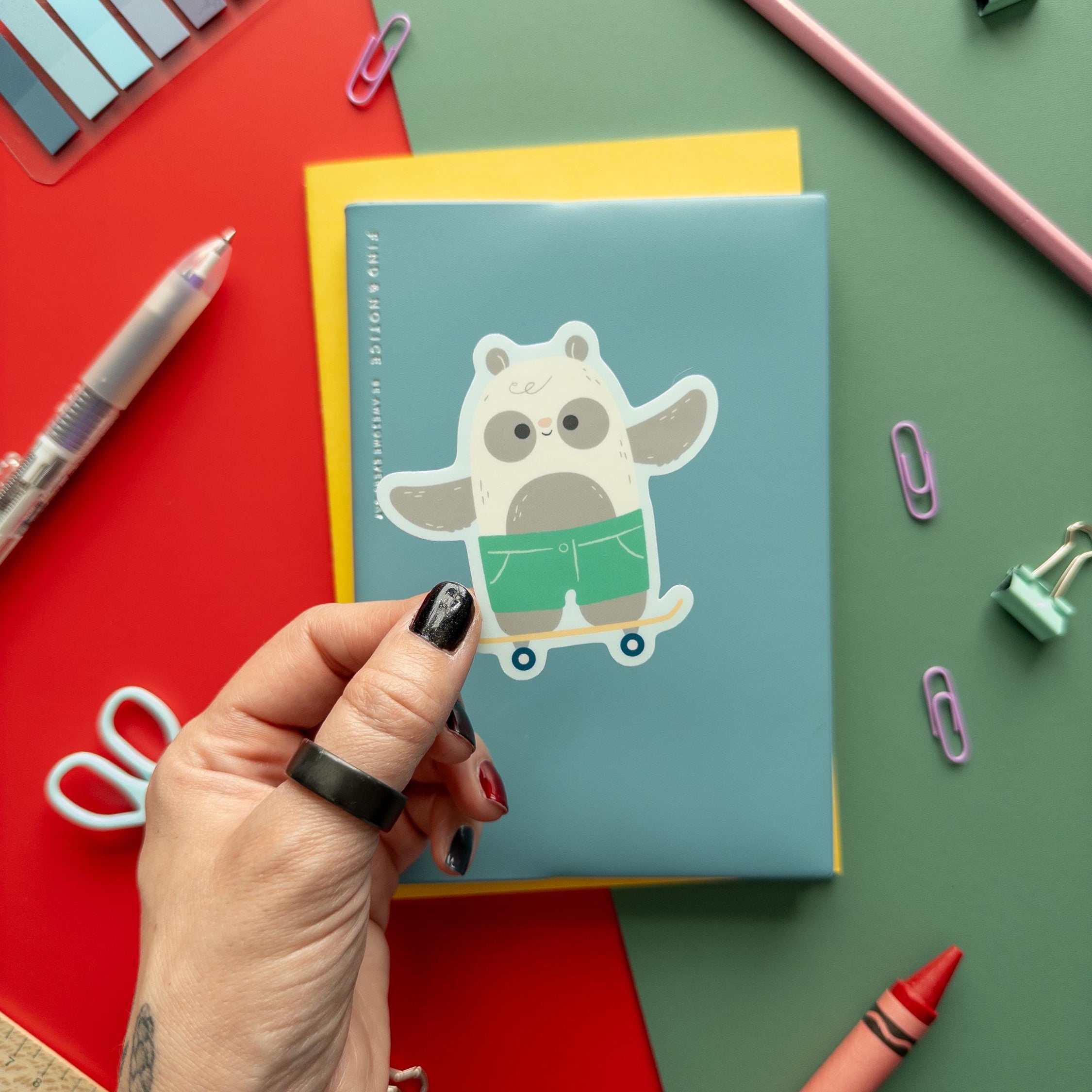 Hand holding a cute skateboarding panda vinyl sticker, waterproof matte decal for laptops, journals, and water bottles, kawaii animal skater design.