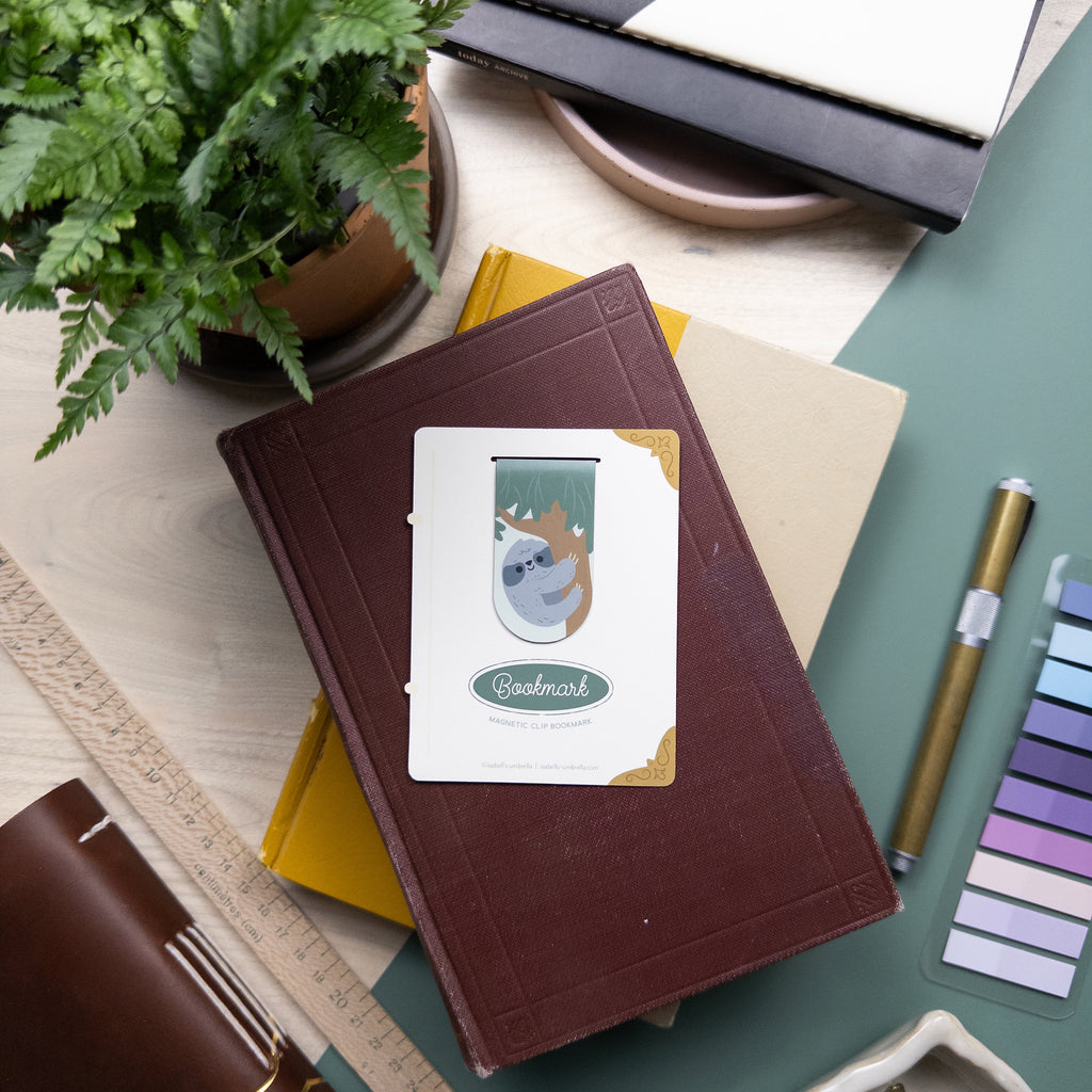 Packaged magnetic clip bookmark featuring an illustration of a sloth pictured on top a stack of books surrounded by a variety of art supplies.