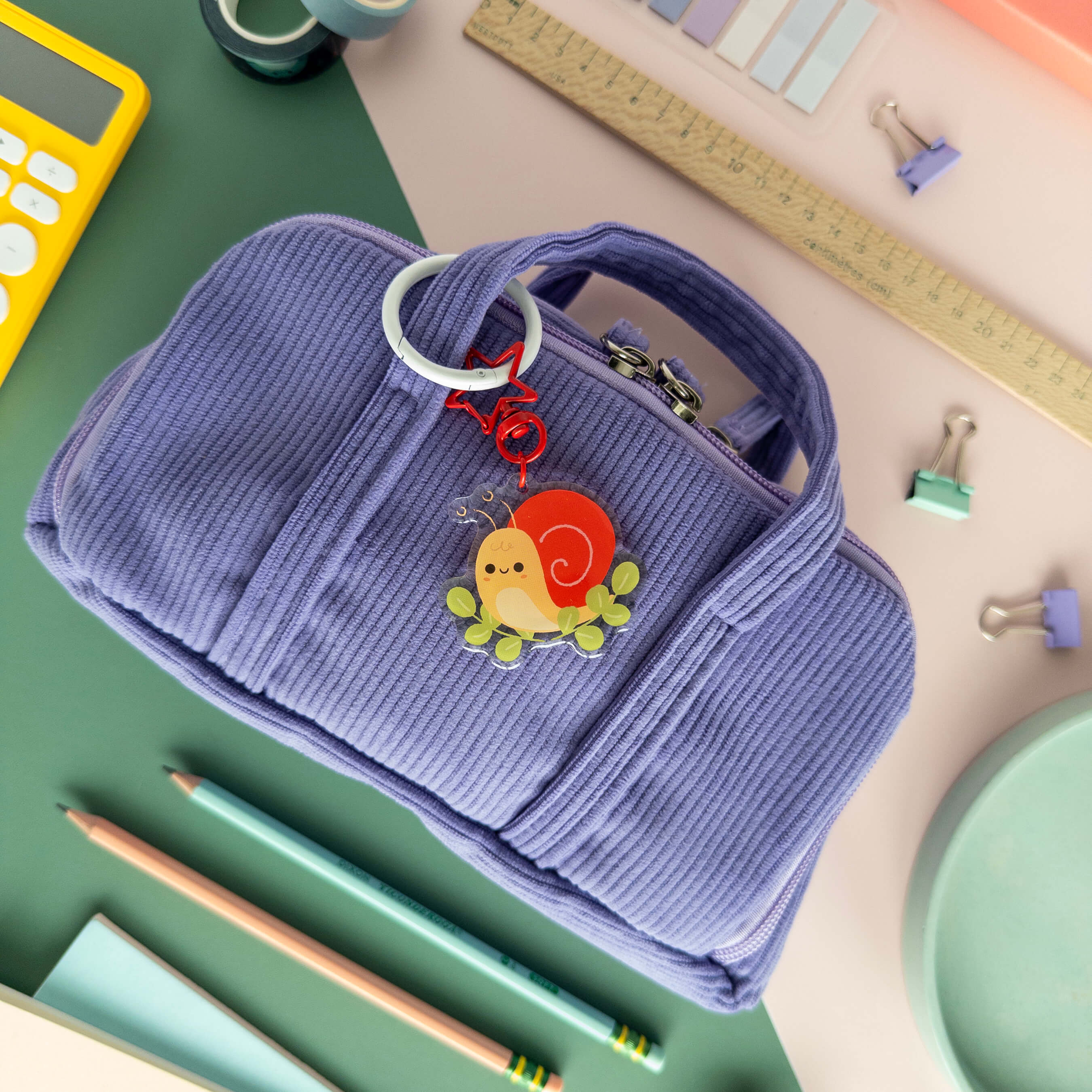 Purple pencil pouch with a keychain of a snail on a desk with stationery items