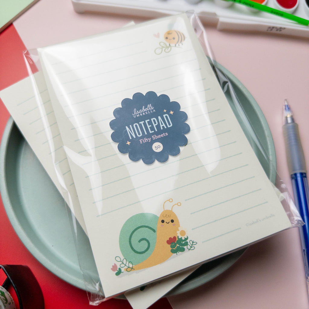Close-up of a cute snail and bee-themed notepad from Isabell’s Umbrella, packaged in clear wrapping with a blue label that reads “Notepad – Fifty Sheets.” The notepad features lined pages with an illustrated snail holding flowers at the bottom and a bee near the top.