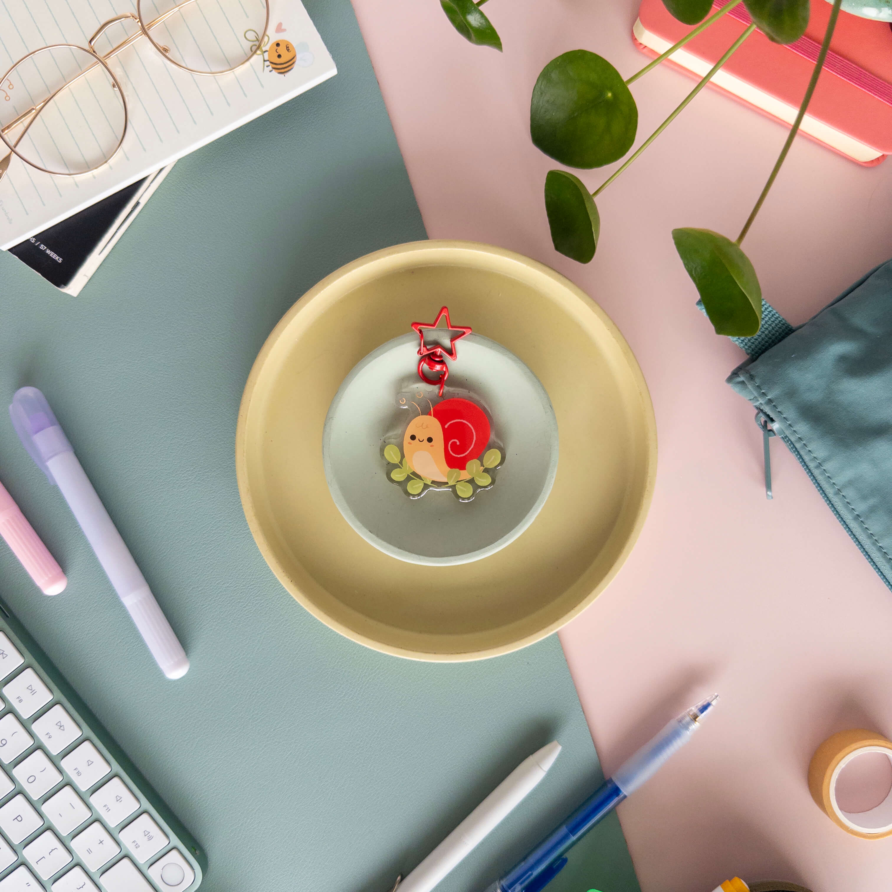 Desk setup with a yellow bowl containing an acrylic snail charm surrounded by pens and a keyboard 
