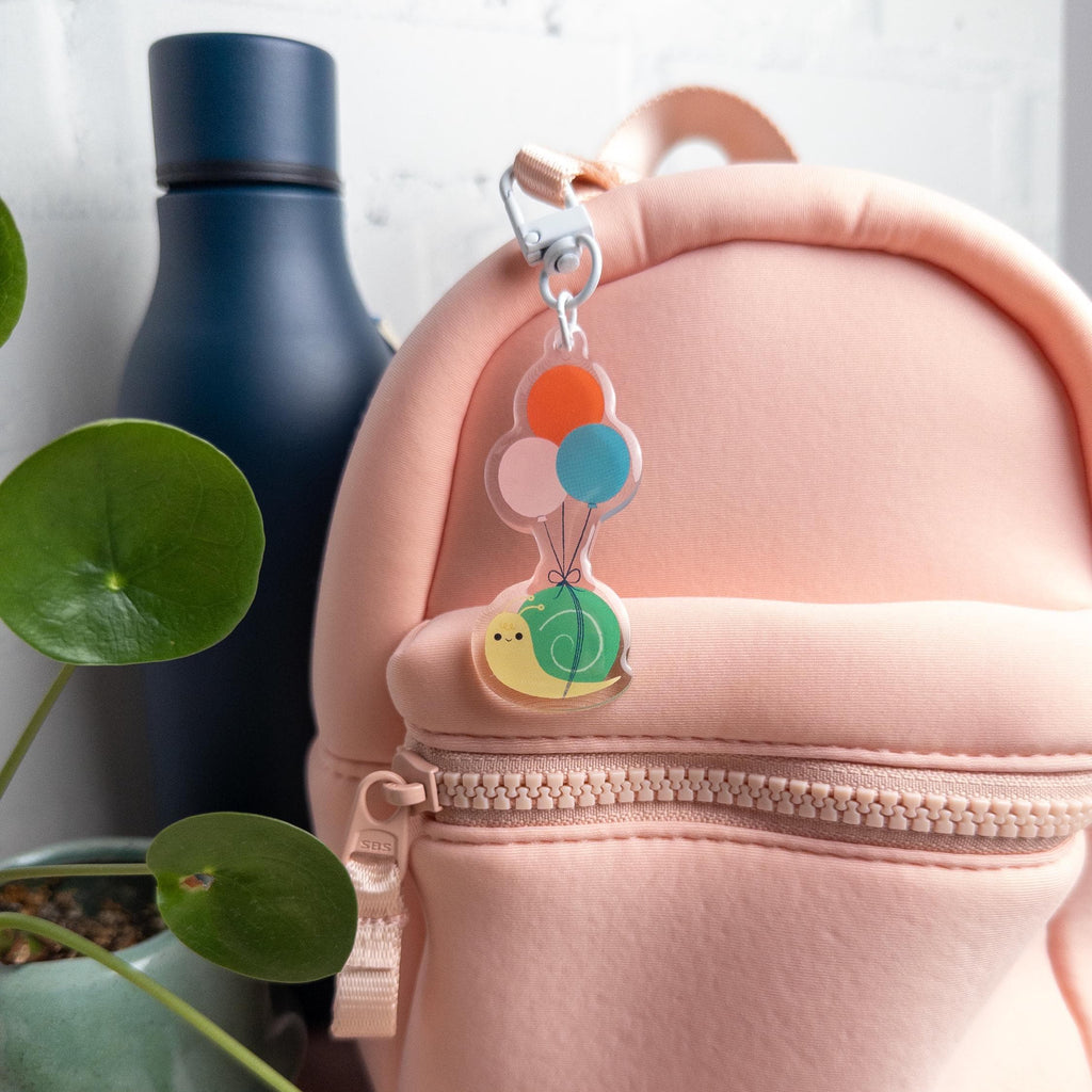Pink backpack with a colorful keychain featuring a snail being carried by balloons attached to its zipper. It is pictured next to a blue water bottle and plant on a light background.