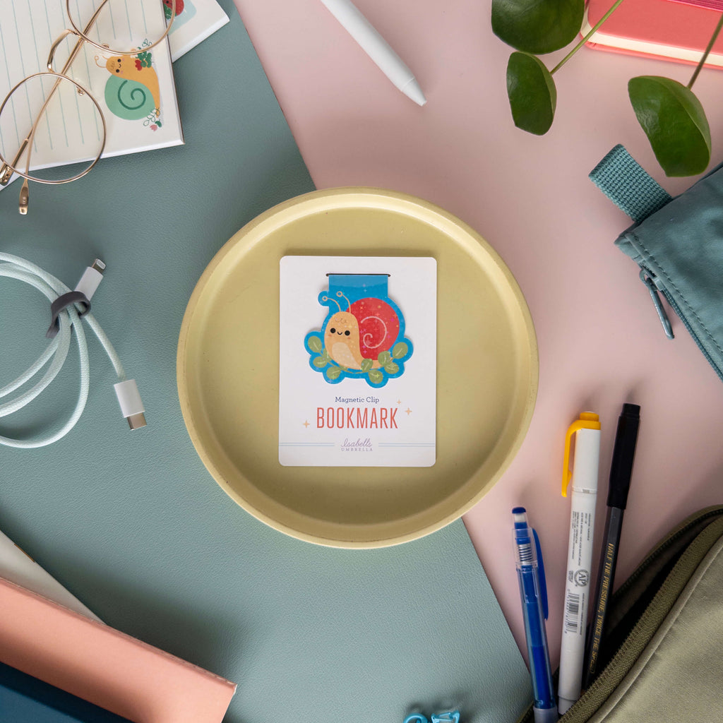 Magnetic snail bookmark on a yellow plate surrounded by stationery items on a colorful background