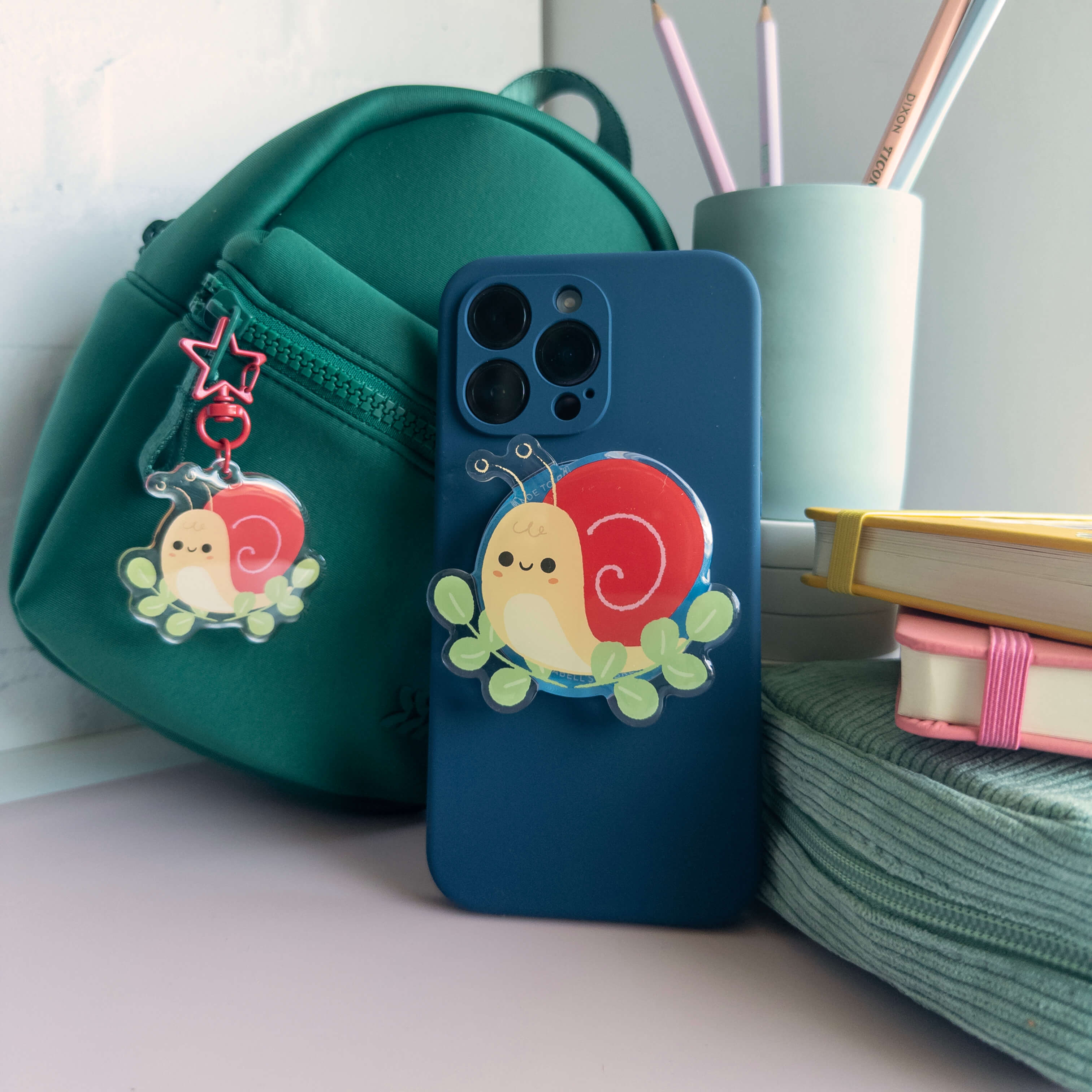 Blue phone case with a cute yellow snail magnetic phone grip, green backpack, and stationery items on a desk.