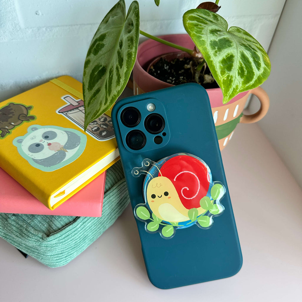 Phone case with a magnetic phone grip of a snail on a surface with books and a plant.