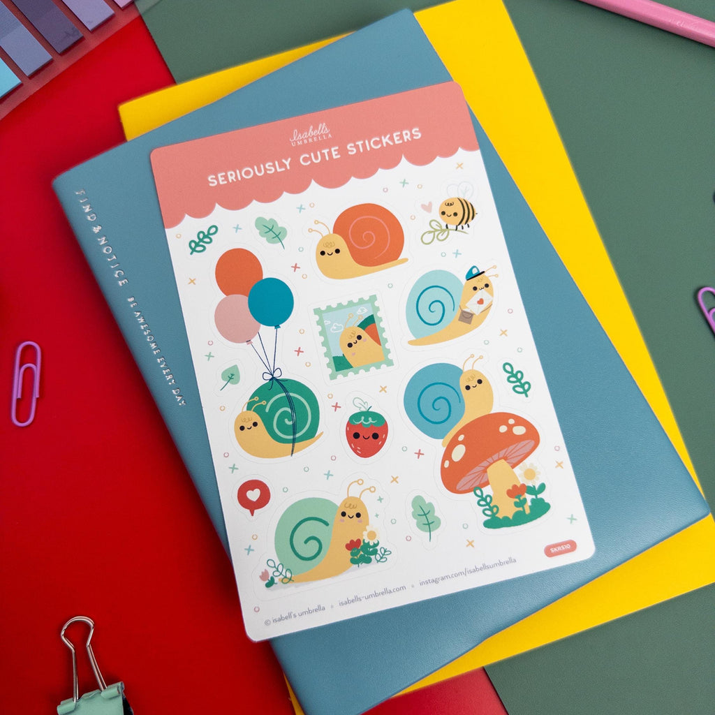 Kawaii snail-themed vinyl sticker sheet by Isabell’s Umbrella placed on a blue and yellow notebook, surrounded by colorful stationery and art supplies.