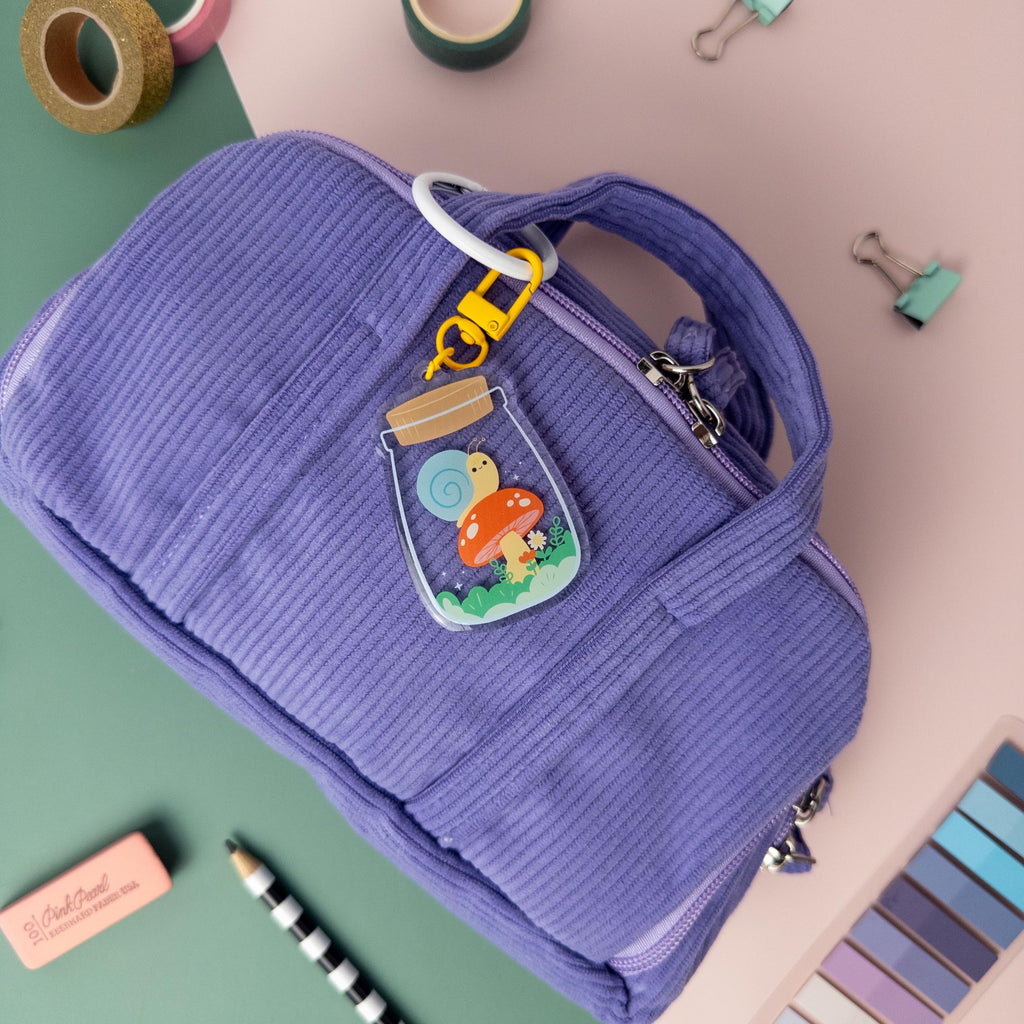 A cute cottagecore snail terrarium acrylic keychain  attached to purple bag pictured on top of a pink and teal background.