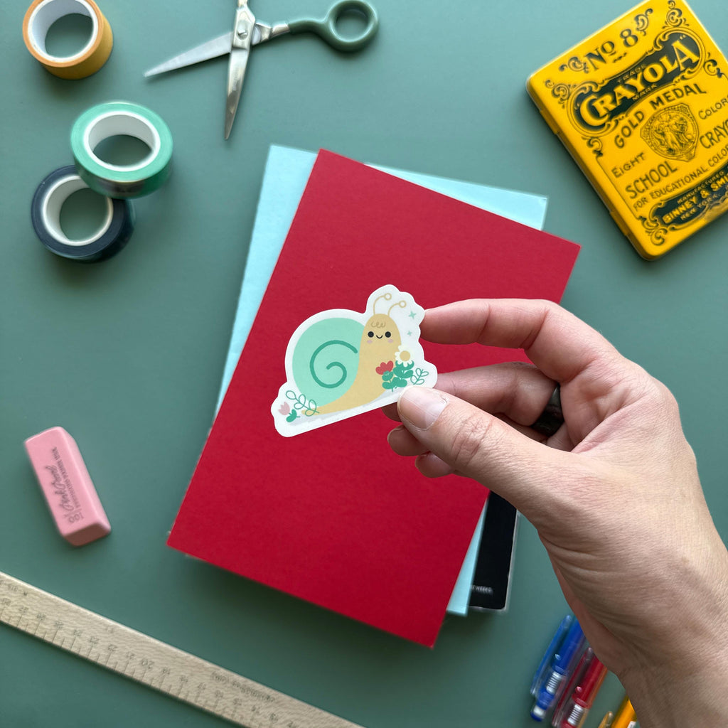 Hand holding a kawaii snail vinyl sticker featuring a sweet snail surrounded by flowers. Surrounded by colorful stationery including washi tape, scissors, and notebooks—ideal for journals, planners, scrapbooking.