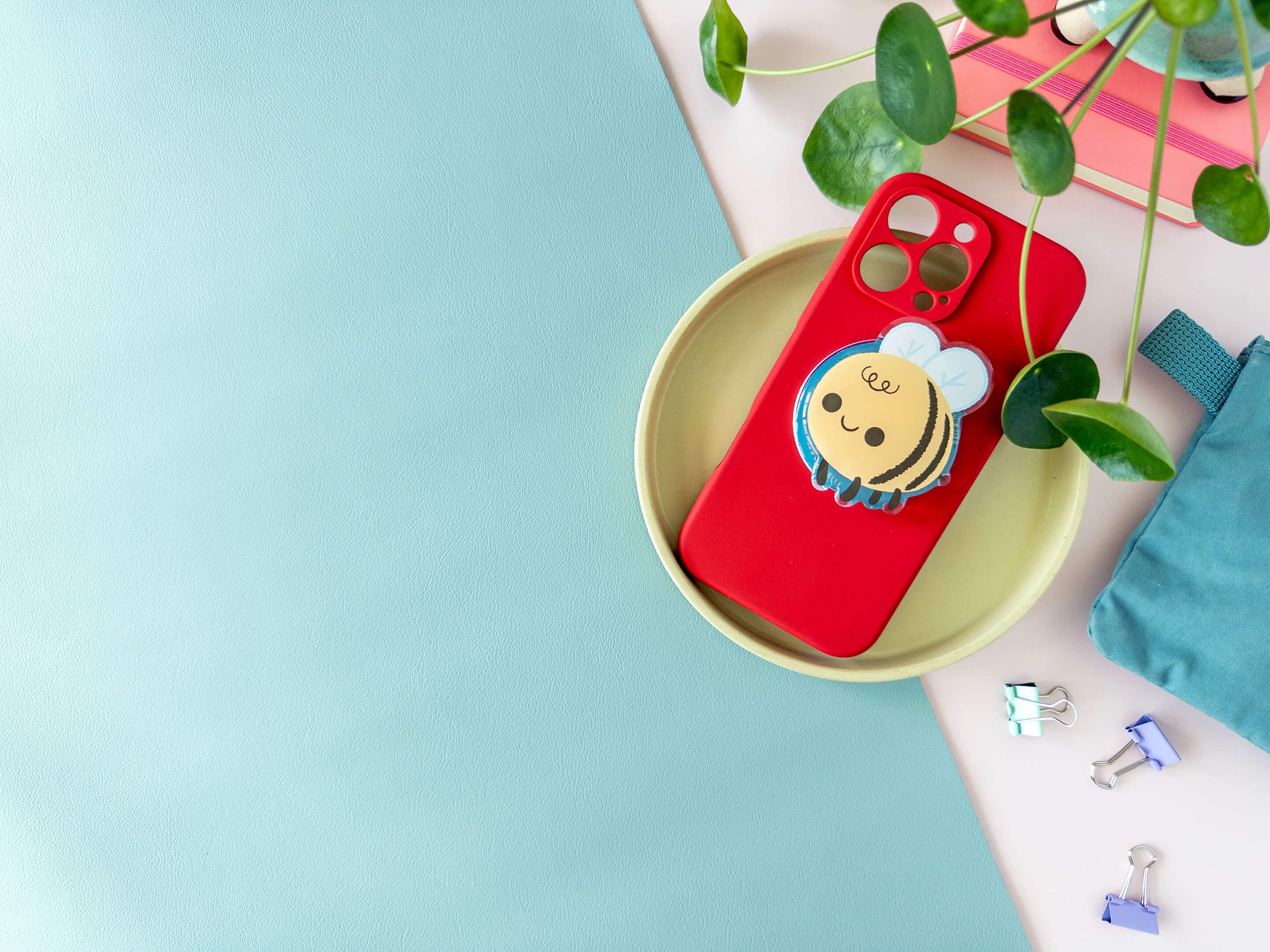 Red phone case with a acrylic bumblebee phone grip on a yellow plate against a light blue and pink background