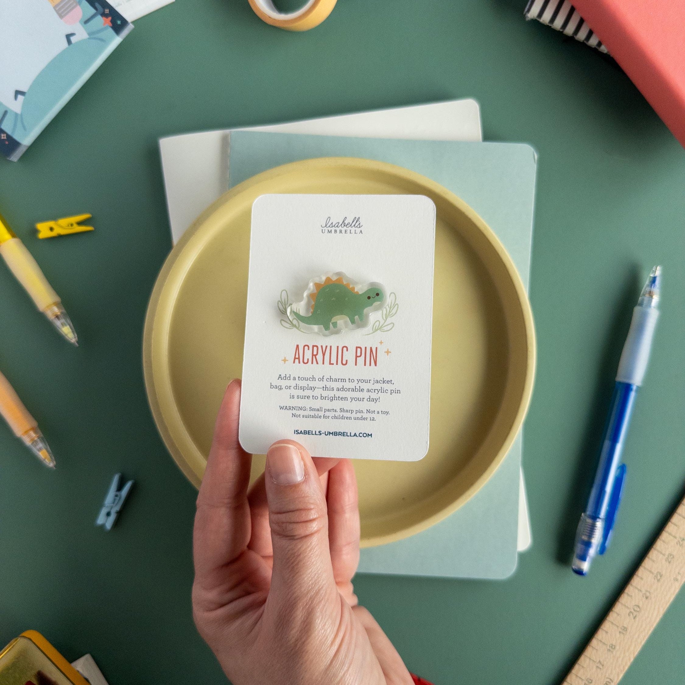 Hand holding an adorable acrylic pin featuring a little green stegosaurus. It is pictured with a variety of art supplies sitting on a green table top.