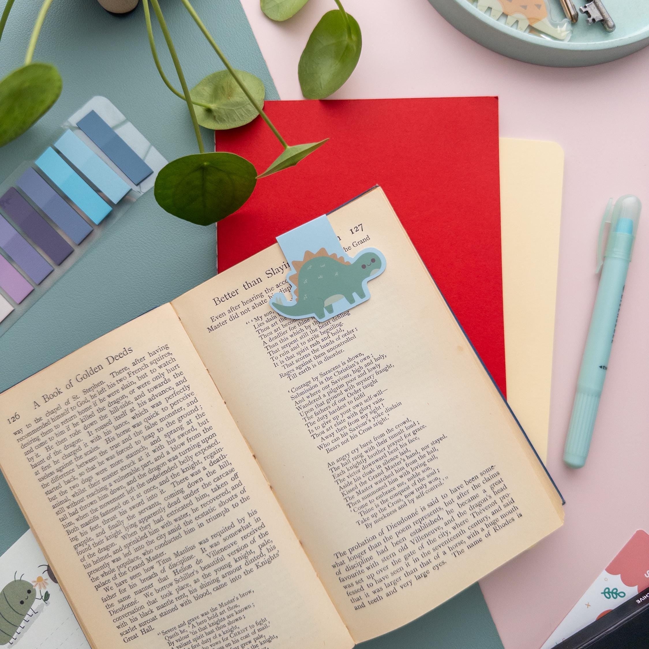 Stegosaurus magnetic bookmark holding a page in an open book, cute dinosaur reading accessory for students and dino fans.