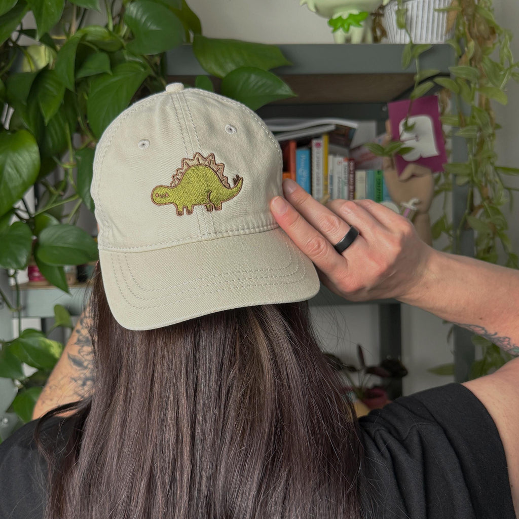 Woman with her back turned to camera wearing a cream cotton twill dad hat with embroidered green stegosaurus design, adjustable dinosaur baseball cap for dino lovers and minimalist fashion fans.