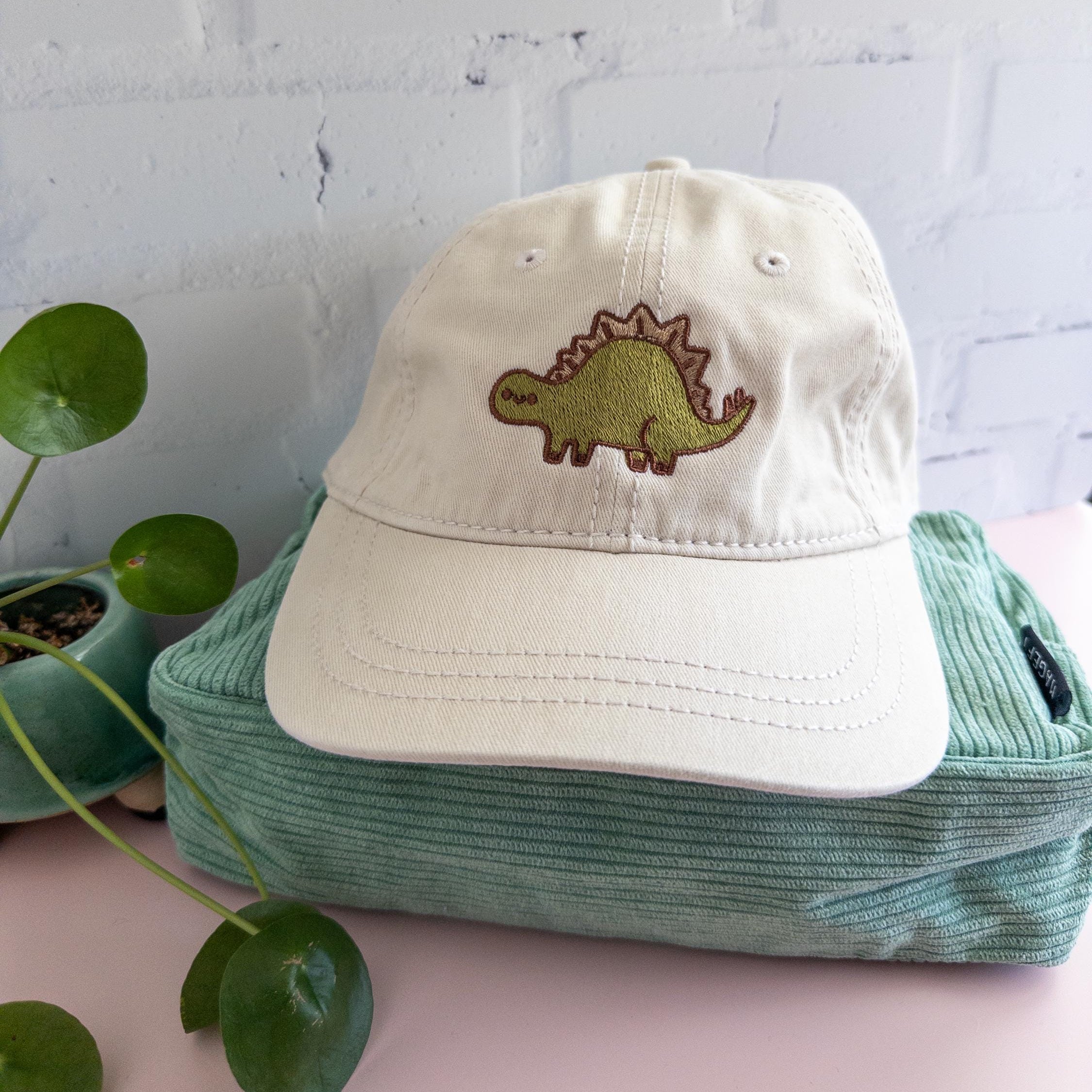 Cream cotton twill dad hat with embroidered green stegosaurus design, adjustable dinosaur baseball cap for dino lovers and minimalist fashion fans. It is photographed on top of a sage green corduroy bag. 