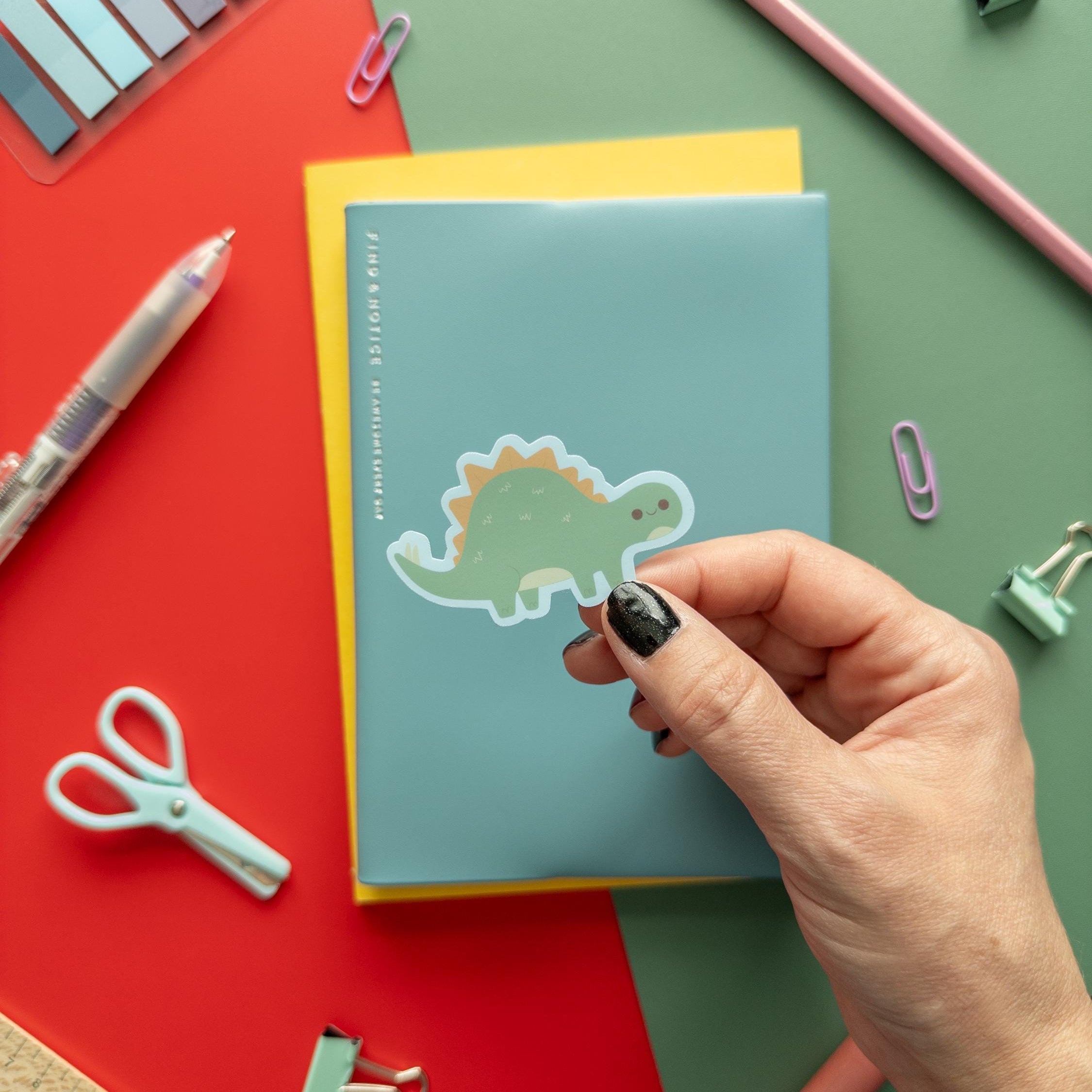 Hand placing a dinosaur sticker on a notebook with stationery items around