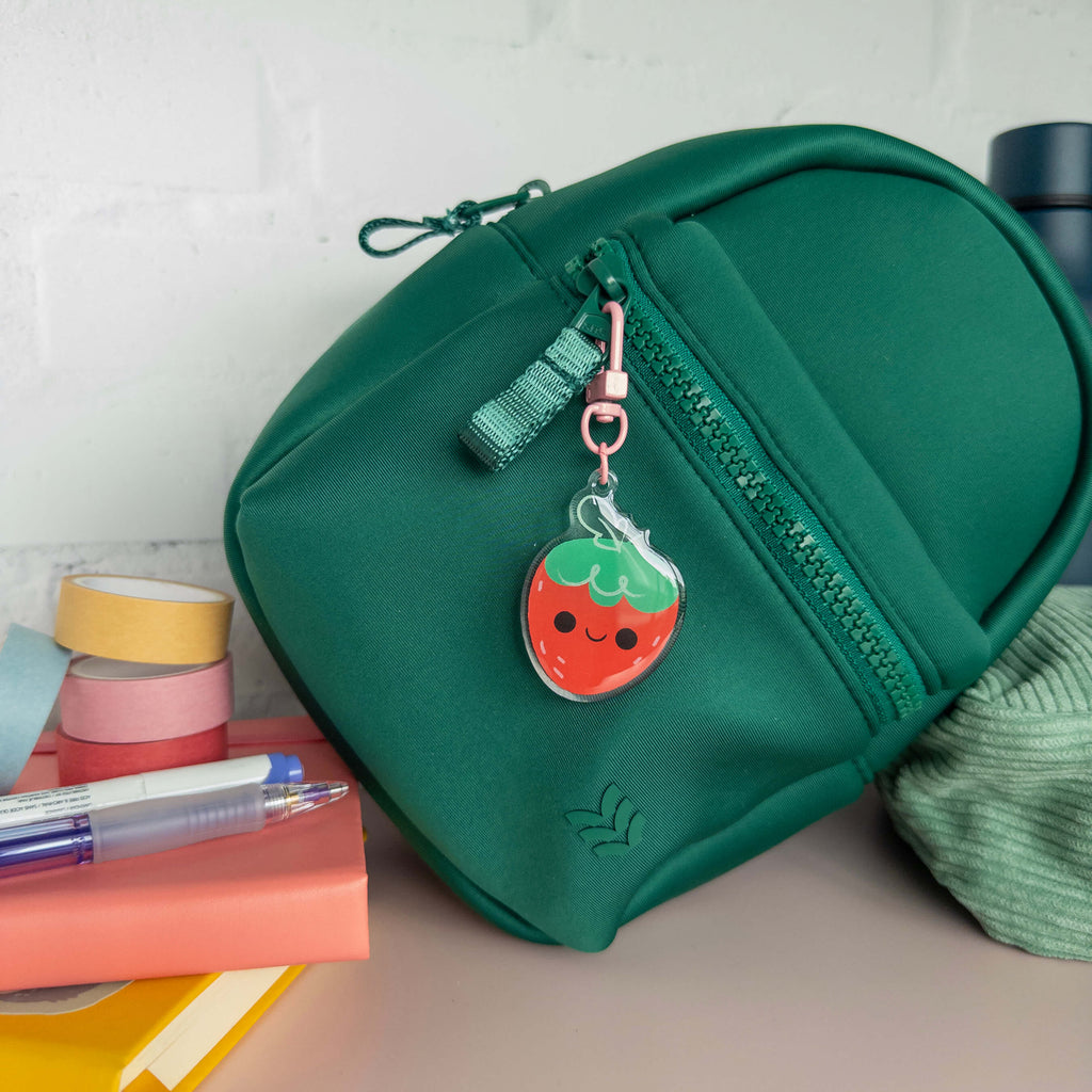 Small green backpack with a strawberry charm on a surface with stationery items