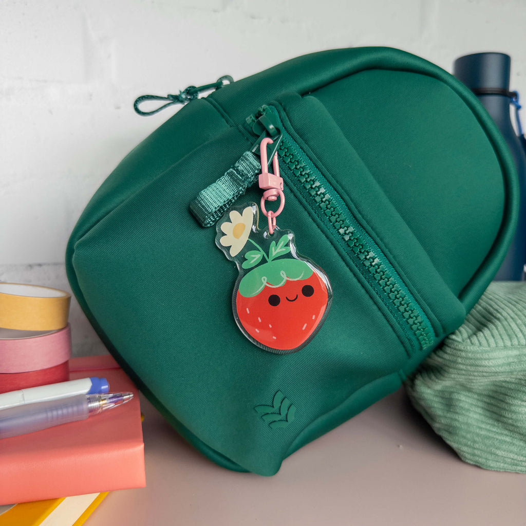 Small green backpack with a strawberry-shaped keychain on a surface with books and a water bottle in the background.
