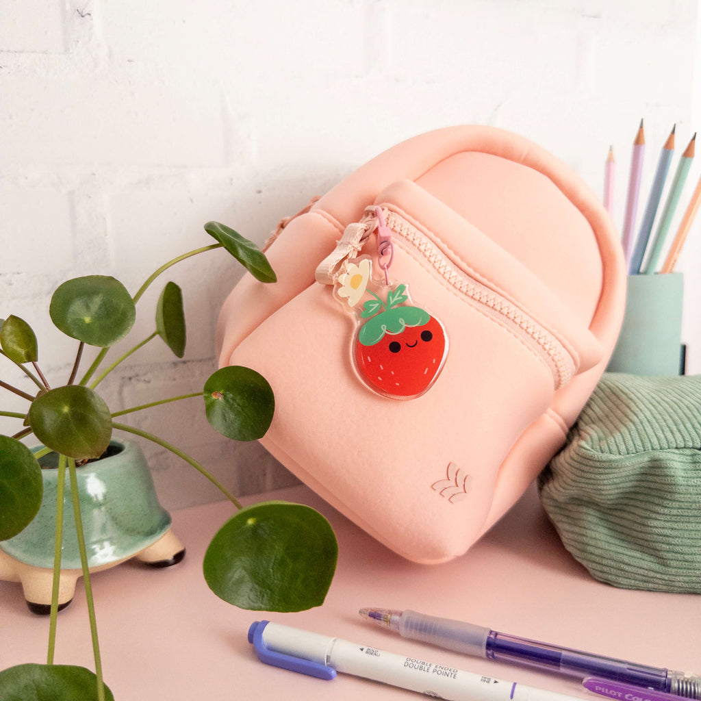 Small pink backpack with a strawberry charm on a desk with stationery items