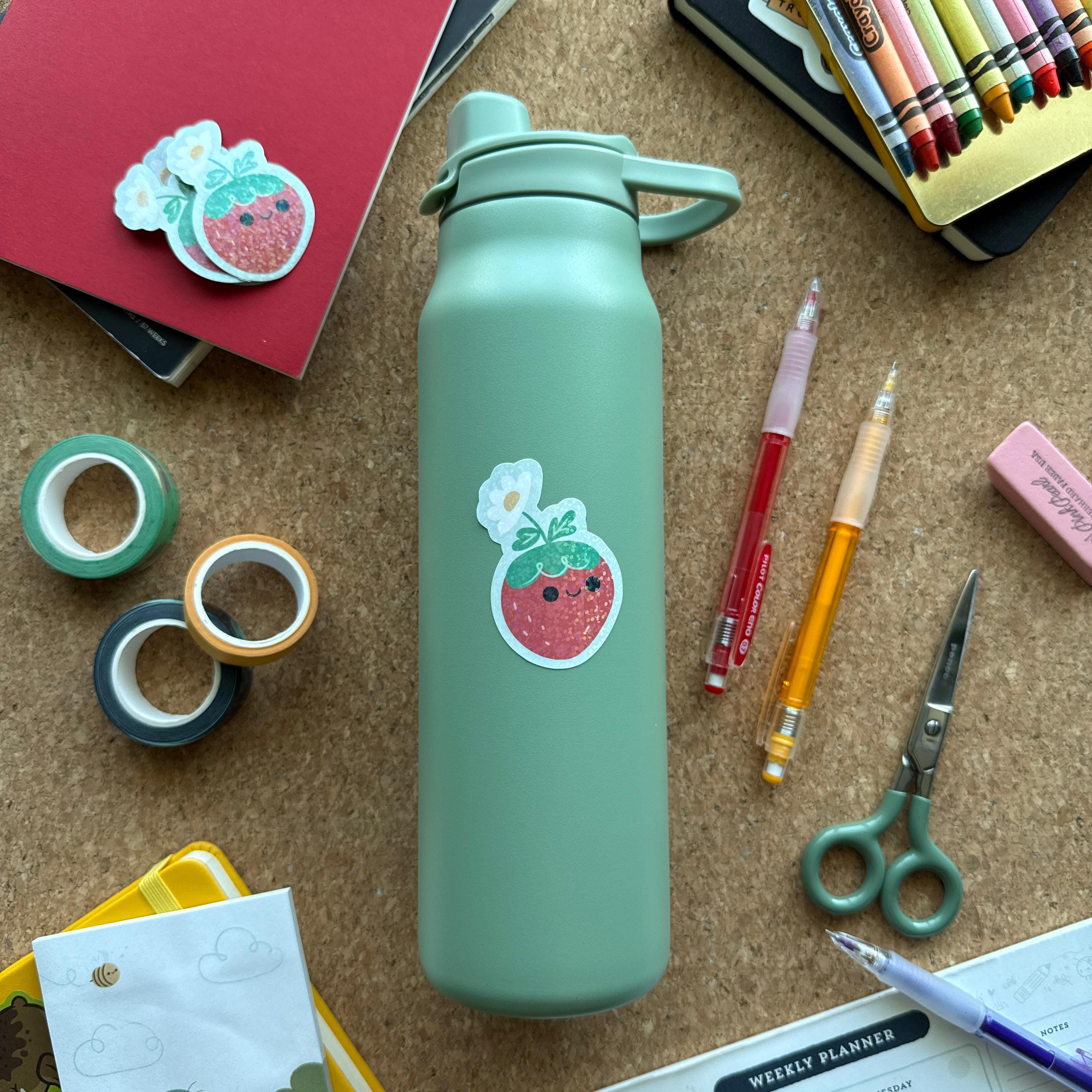 Cute strawberry with a flower glitter sticker on a sage green water bottle, surrounded by colorful pens, washi tape, crayons, and notepads. This waterproof vinyl sticker is perfect for decorating bottles, laptops, and school supplies.