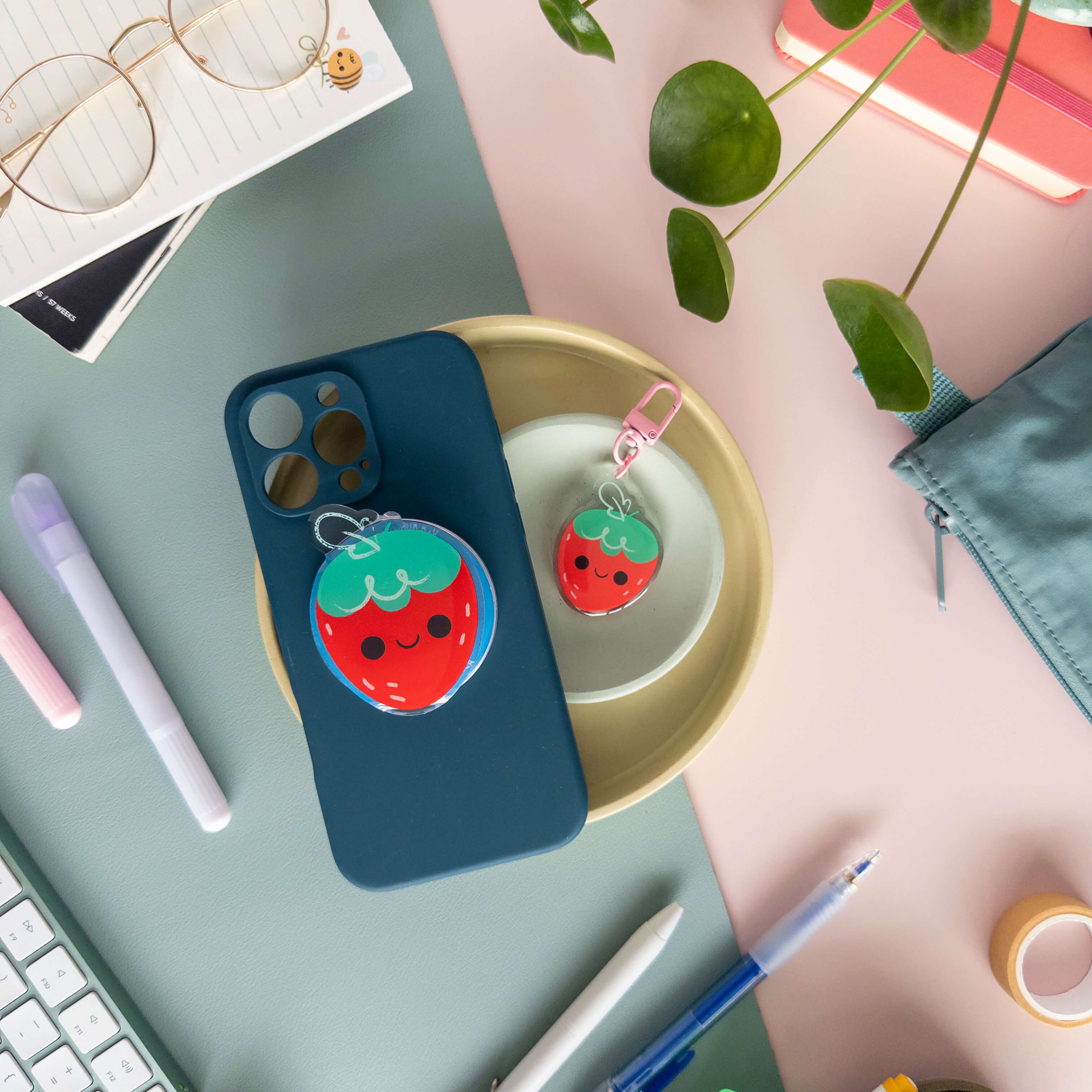 Phone case with strawberry magnetic phone grip next to a strawberry charm in a bowl on a desk with stationery items