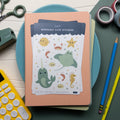 Sticker set of sea related illustrations like a narwhale, sea horse, shrimp, sea urchin and more, laying on top of a stack of notebooks pictured with pencils, washi tape, and other office related items.