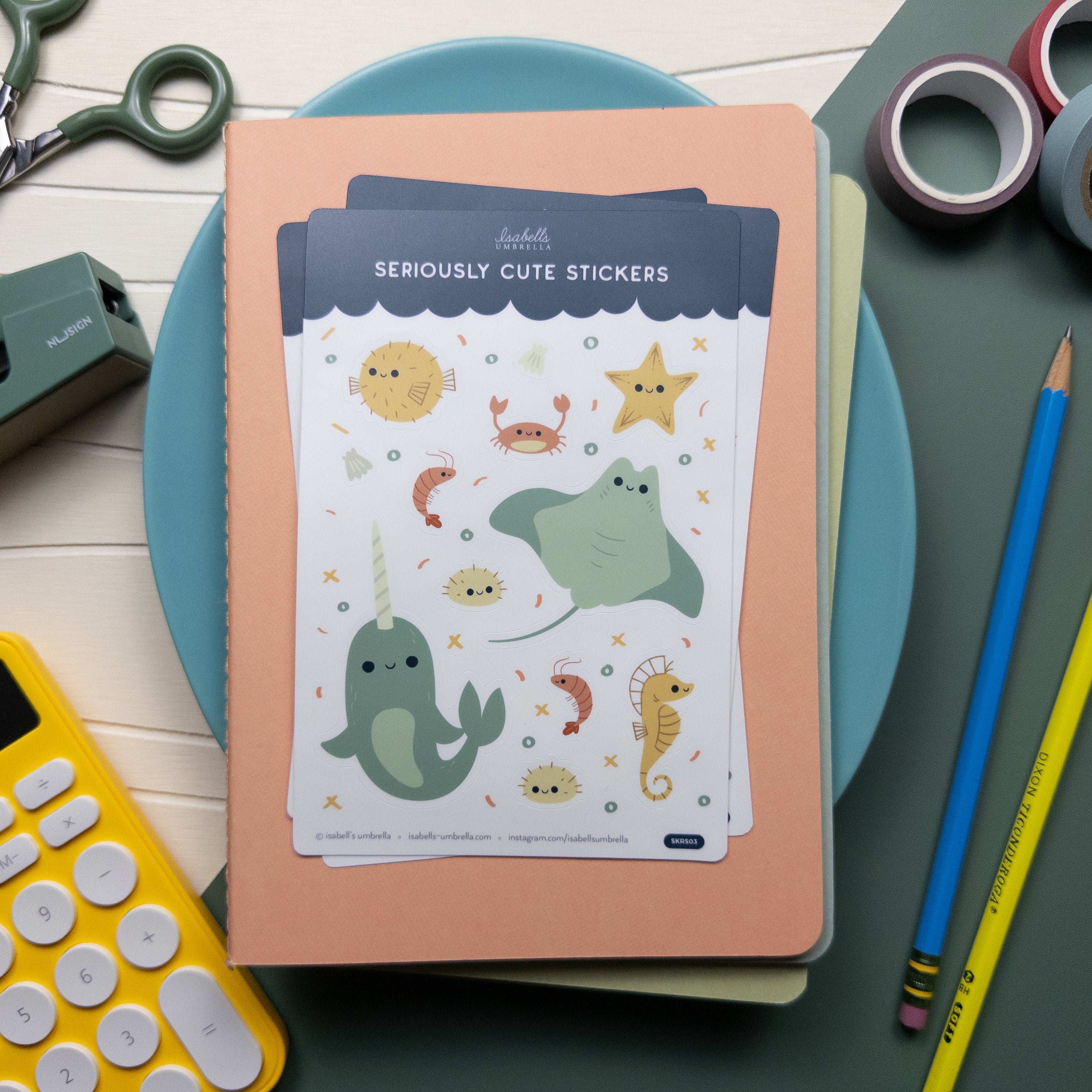 Sticker set of sea related illustrations like a narwhale, sea horse, shrimp, sea urchin and more, laying on top of a stack of notebooks pictured with pencils, washi tape, and other office related items.
