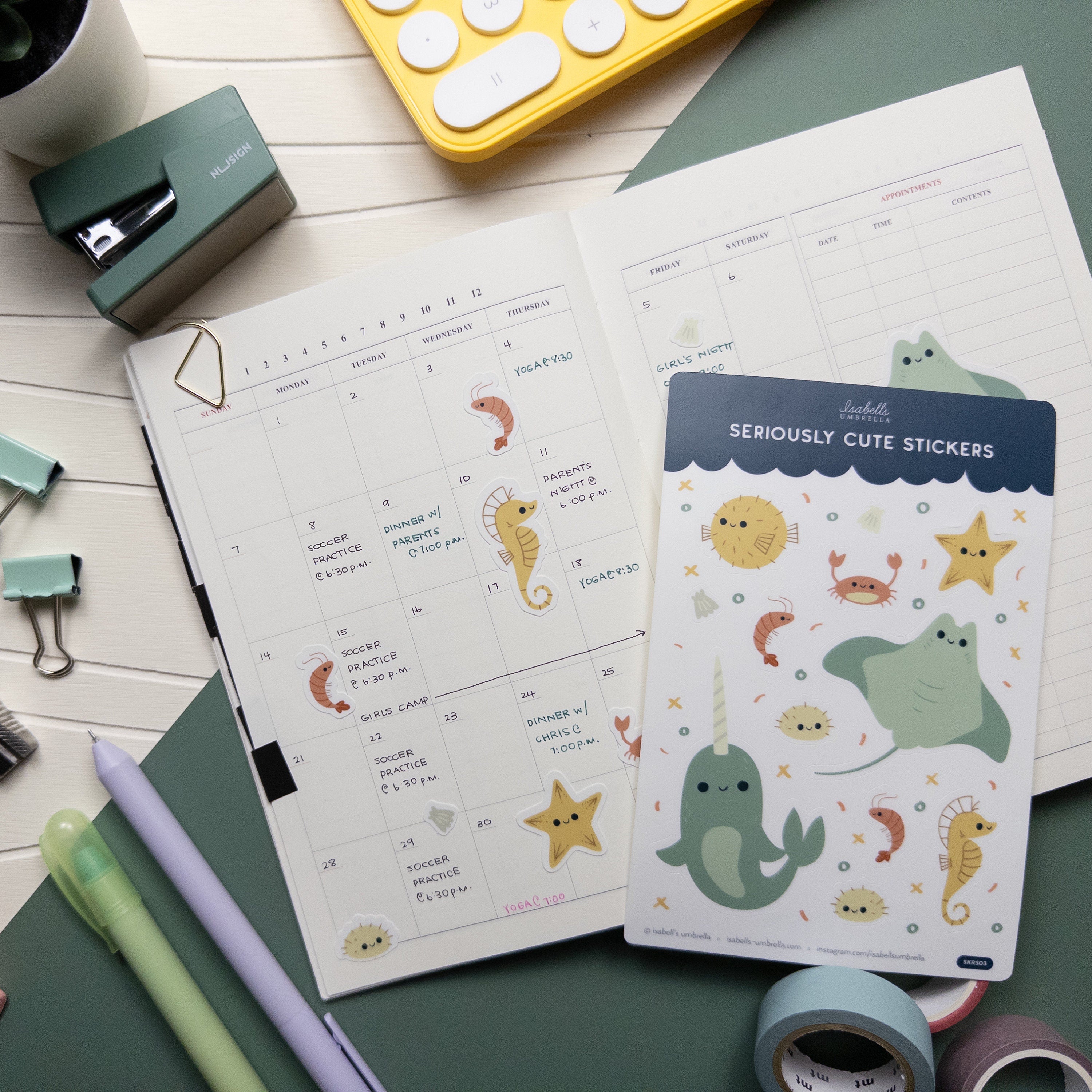 Flatlay of a calendar surrounded by office supplies and a sheet of stickers featuring a theme of sea related creatures.