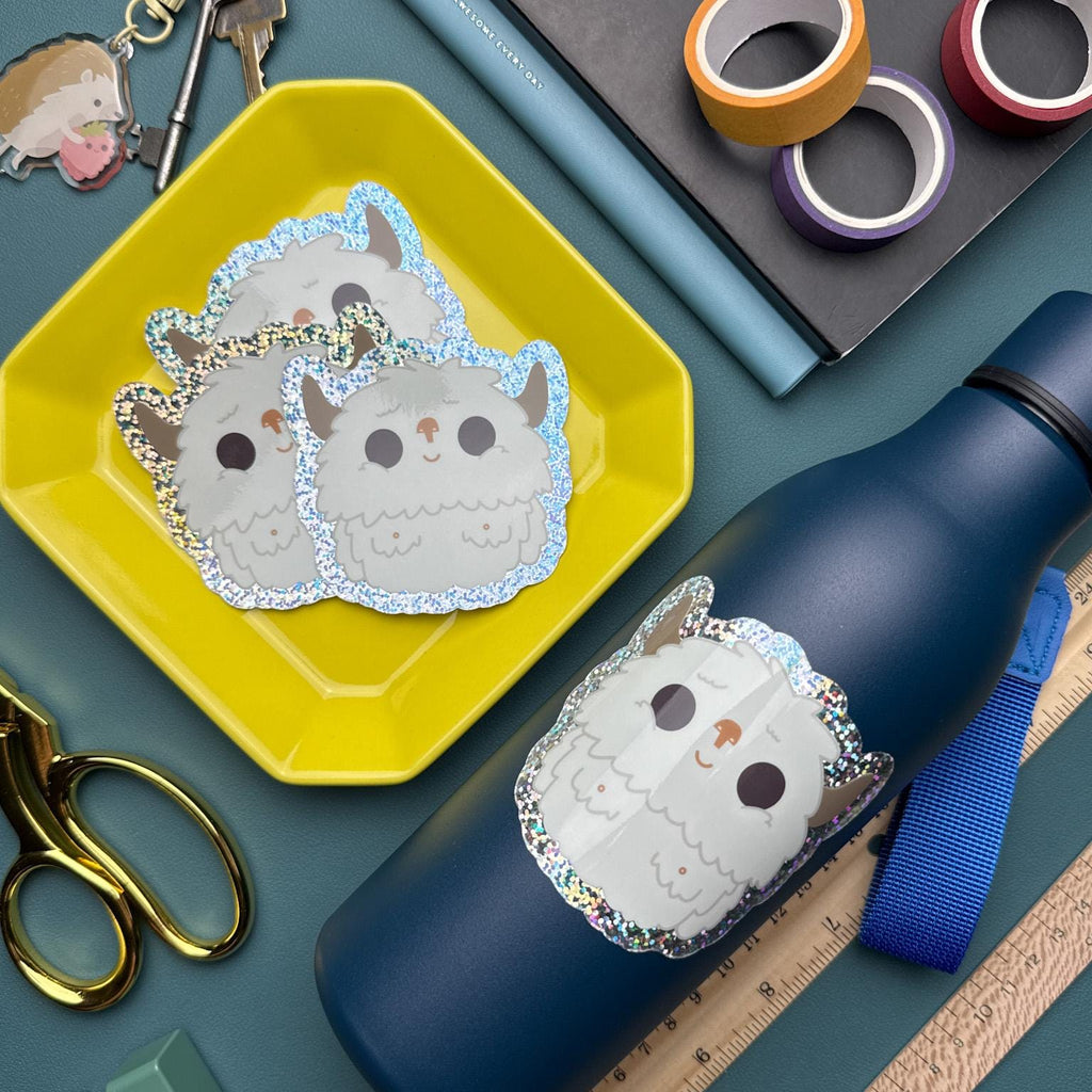 A cute glittery yeti vinyl sticker attached to a blue water bottle sitting next to a yellow plate with a pile of yeti stickers in it. They are surrounded by a variety of office supplies including a ruler, scissors, and washi tape.